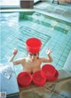 A young boy in a swimming pool with red buckets on his head.